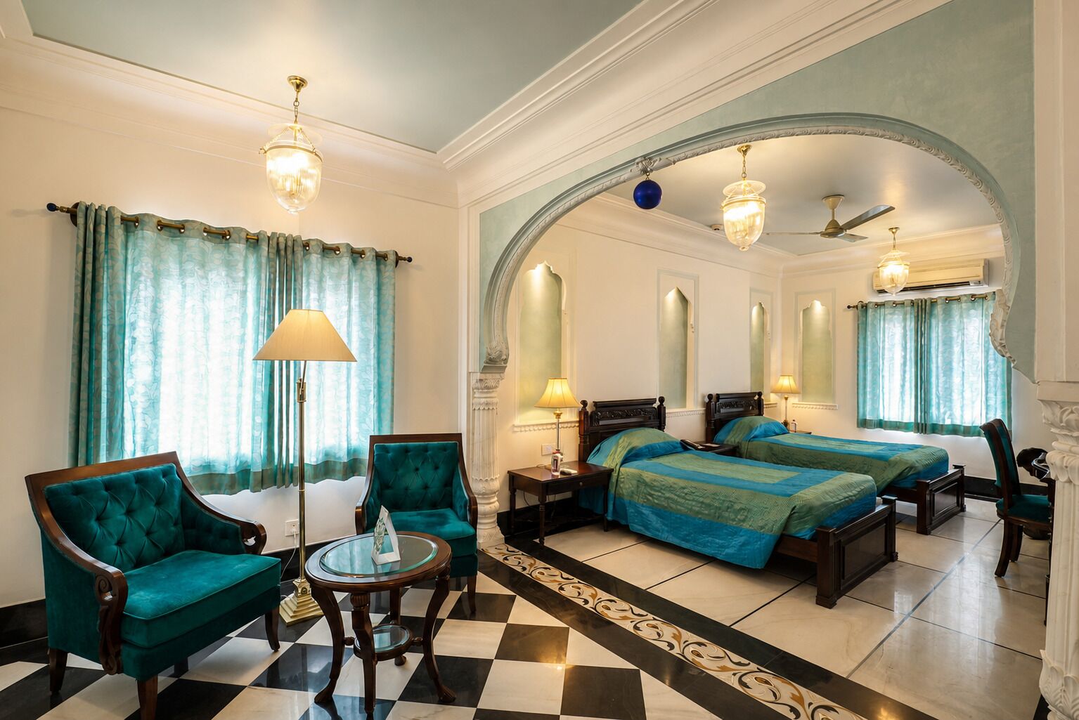 A hotel room with a checkered floor, two beds, a sitting area with a table and chairs, and a large arched doorway.