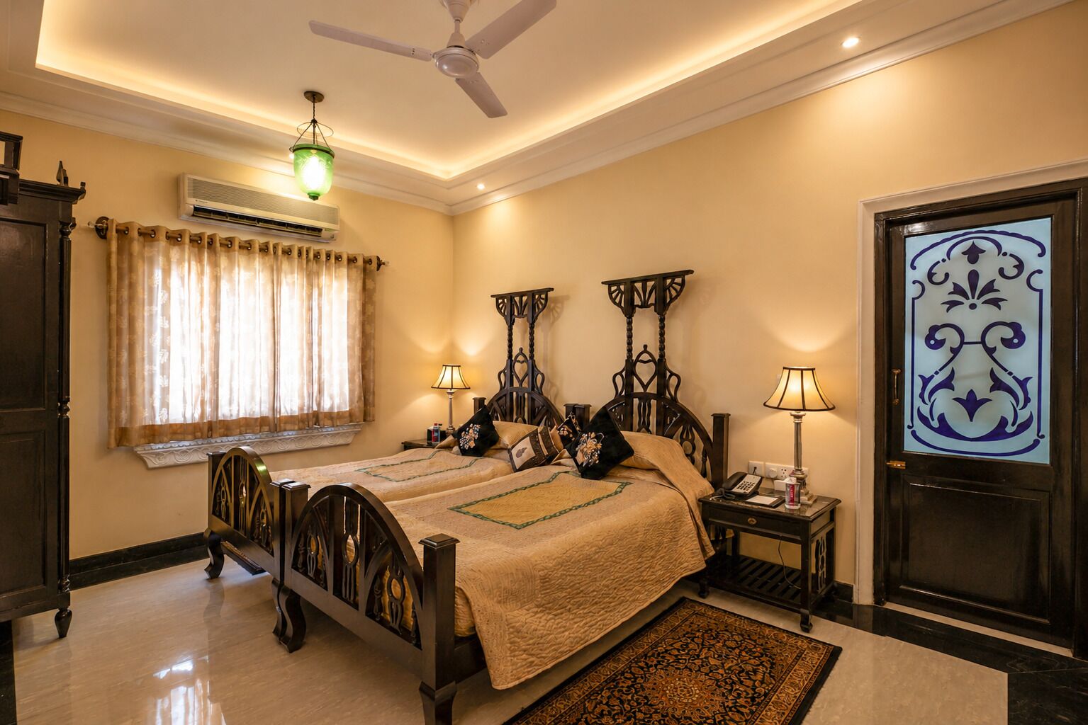 A double bed with a dark wooden frame, two bedside tables with lamps, a ceiling fan, an air conditioning unit, and a decorative door.