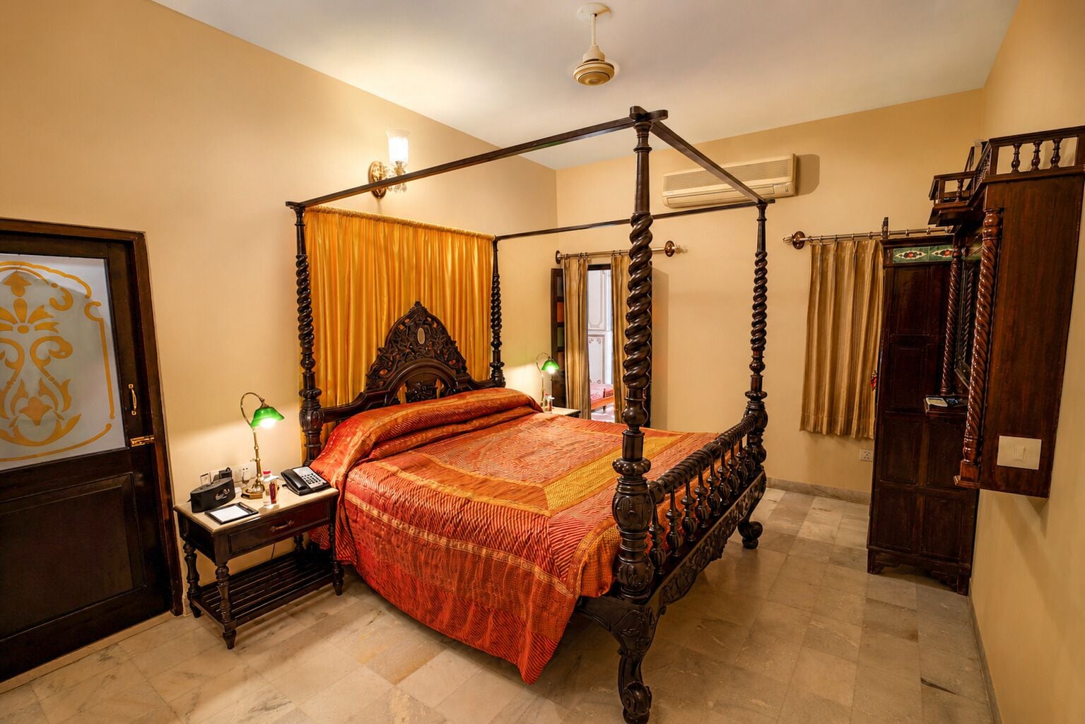 Una cama con dosel de madera y una colcha roja, una mesita de noche con una lámpara, y una puerta con un panel de vidrio decorativo.