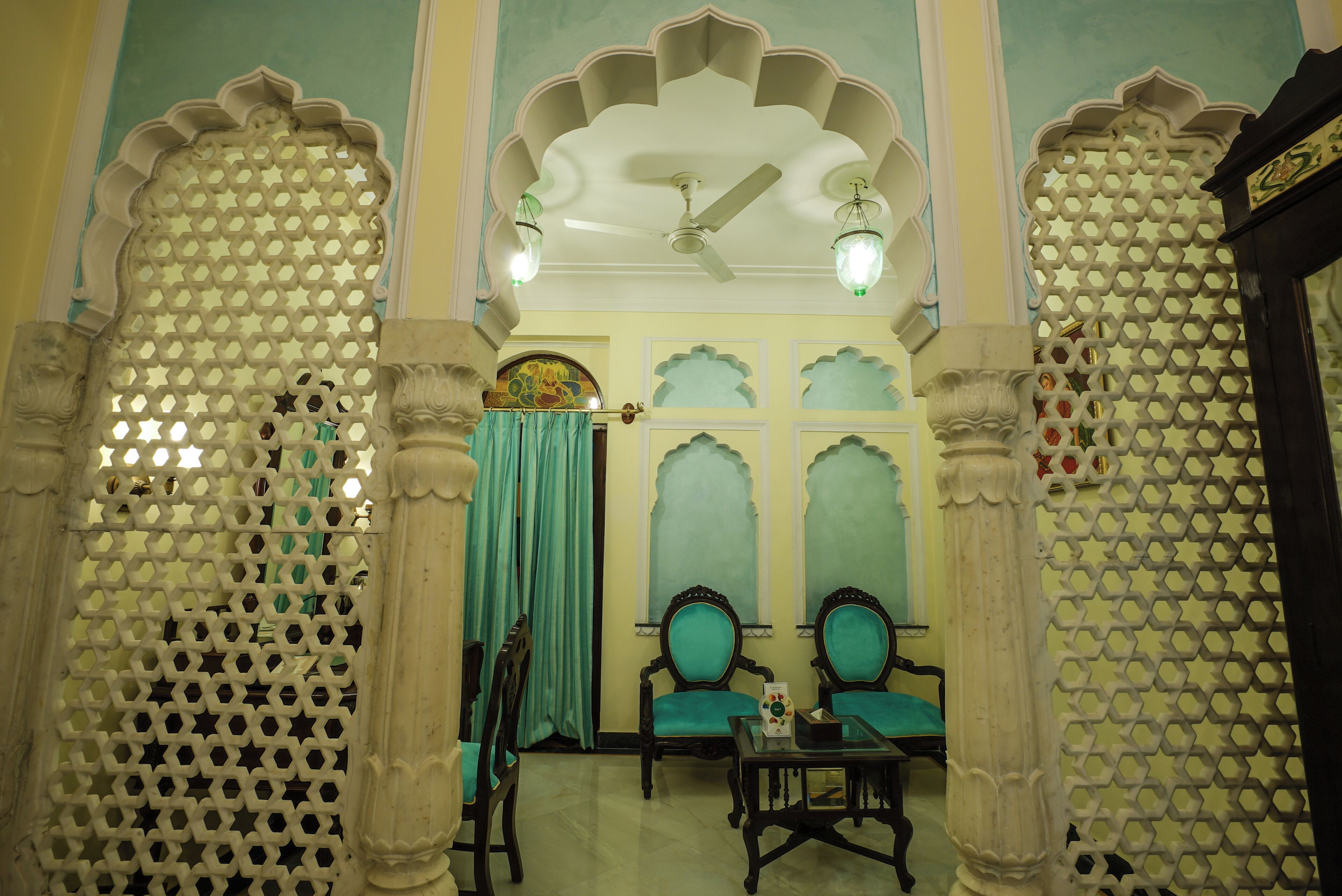 Une pièce dotée d’éléments architecturaux traditionnels, notamment des colonnes décorées et un ventilateur de plafond. On y trouve deux fauteuils avec des coussins et une petite table sur laquelle est posée une boîte.