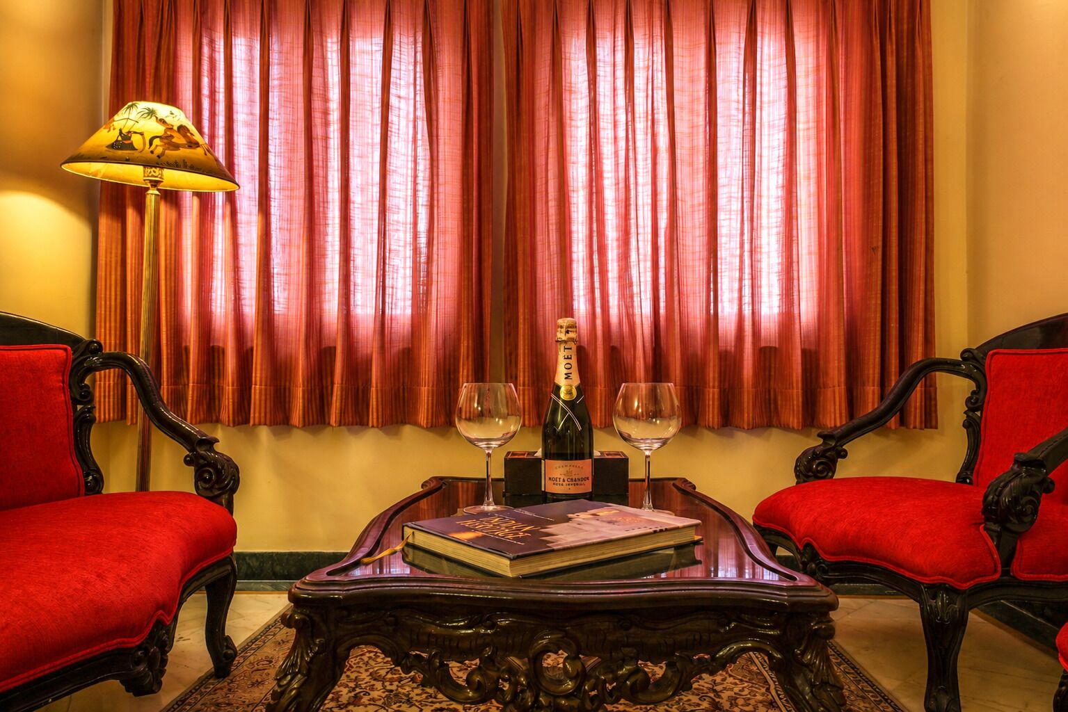 Um quarto com duas poltronas vermelhas, uma mesa de centro de madeira com um livro e uma garrafa de champanhe, e um abajur.