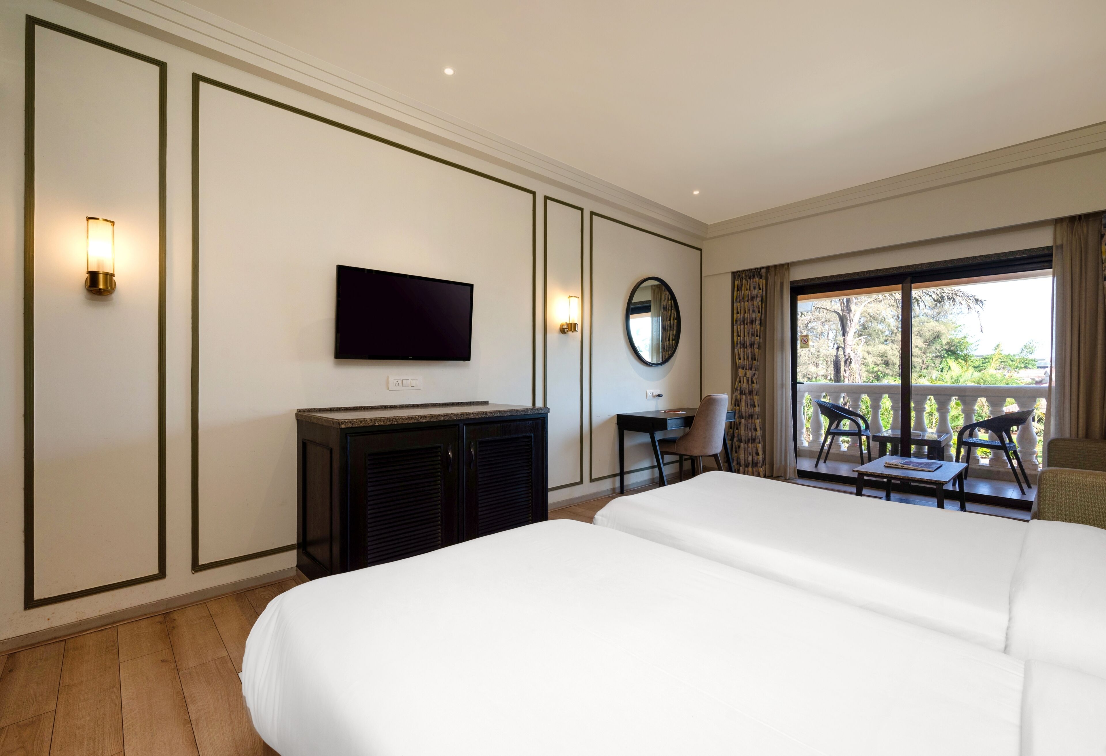 A hotel room with a large bed, a desk with a chair, a television, a mirror, and a balcony with chairs.