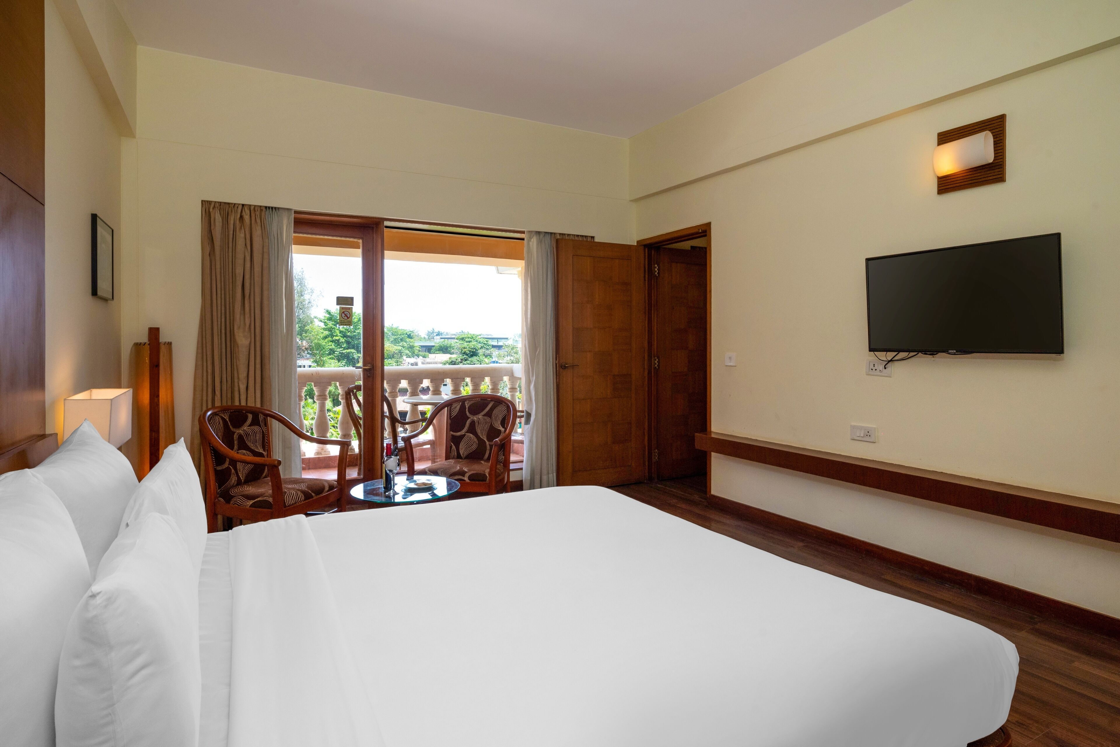 A hotel room with a large bed, a TV mounted on the wall, a balcony with a table and chairs, and a view of greenery outside.