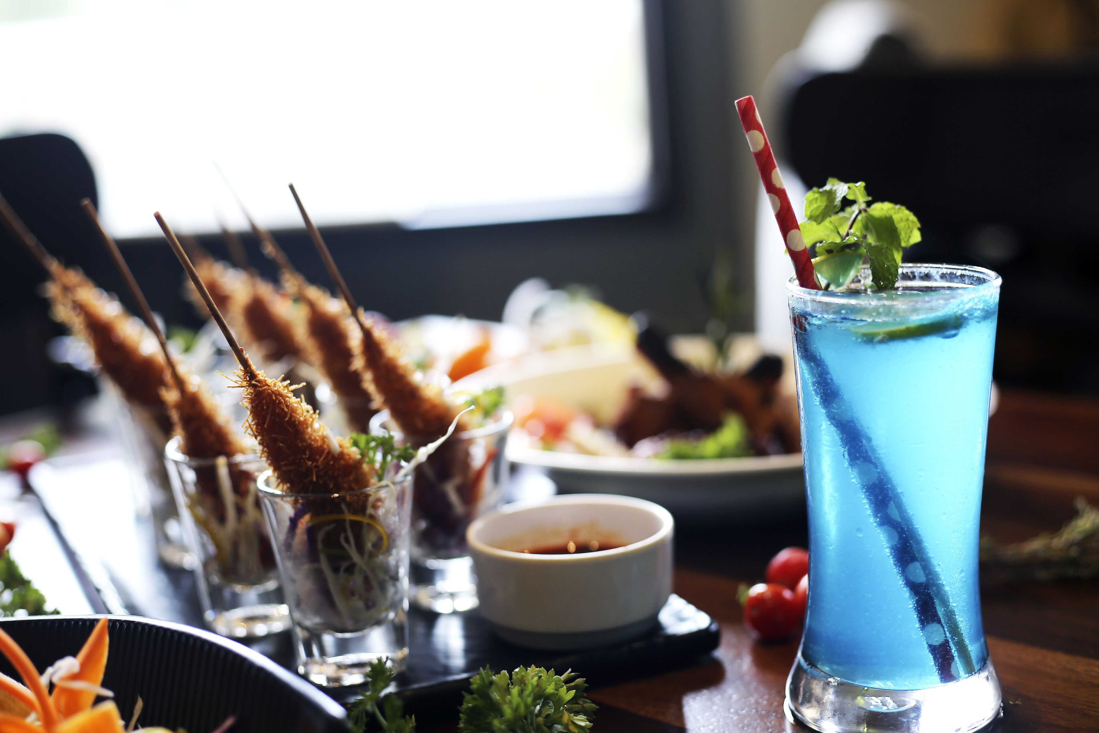 Une table à manger avec des brochettes de nourriture, un verre d’une boisson bleue et un petit bol de sauce.