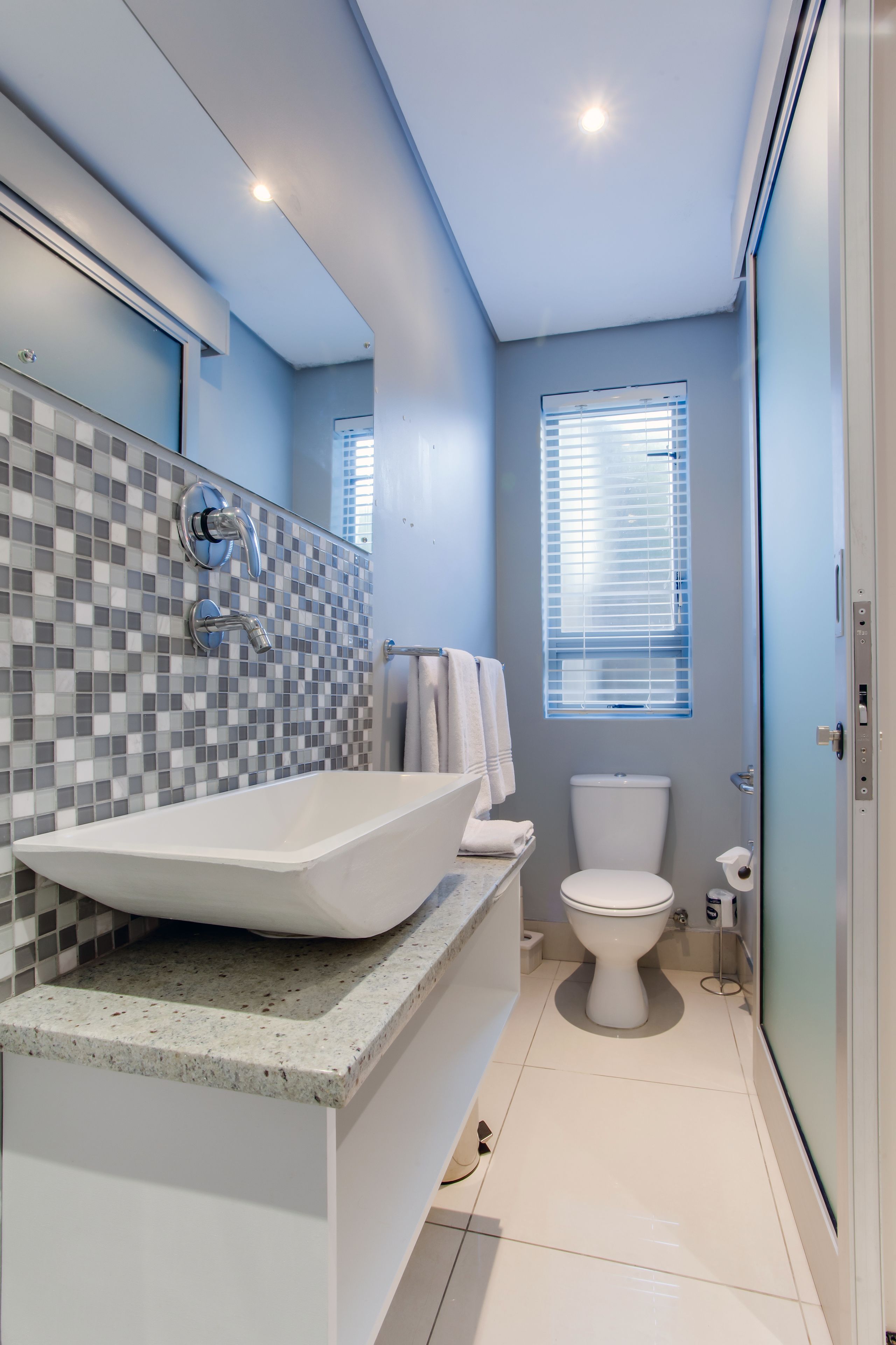 A modern bathroom with a large vessel sink, a toilet, and a window with blinds.