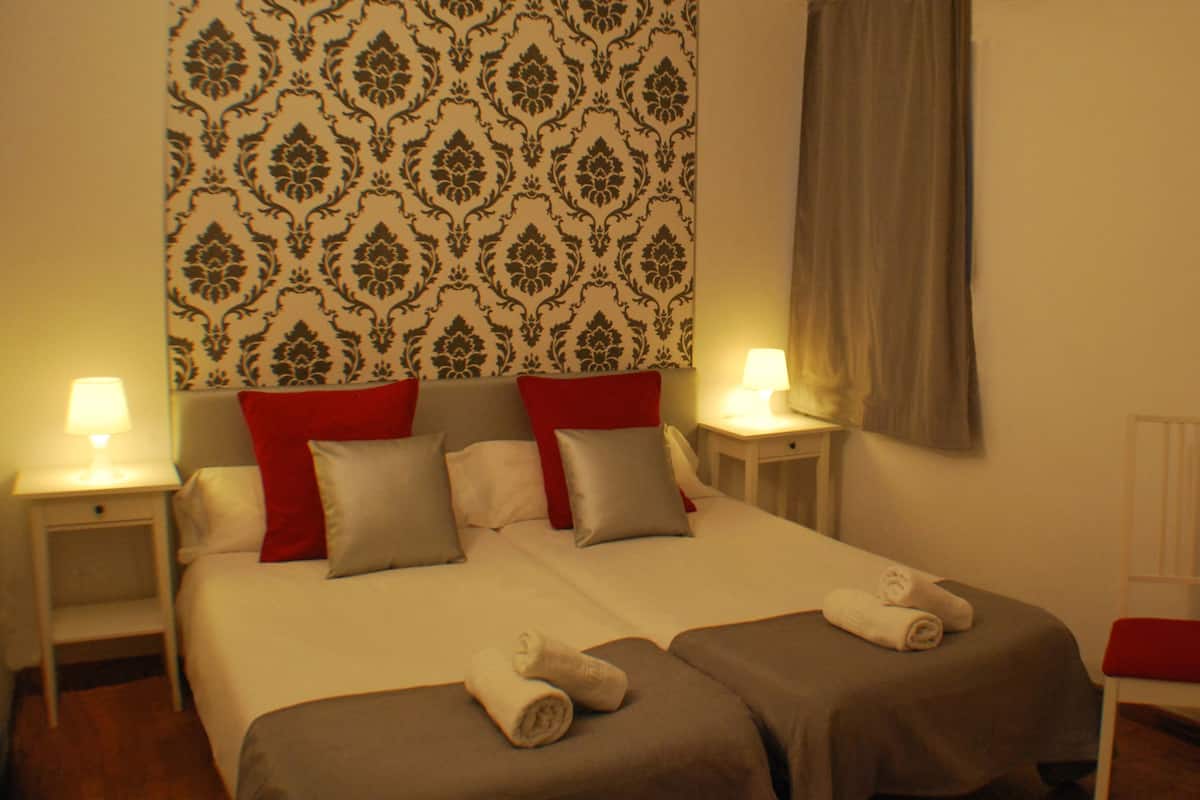 Habitación de hotel con dos camas, papel tapiz rojo y blanco estampado, una silla roja y una mesa pequeña con mantel rojo.