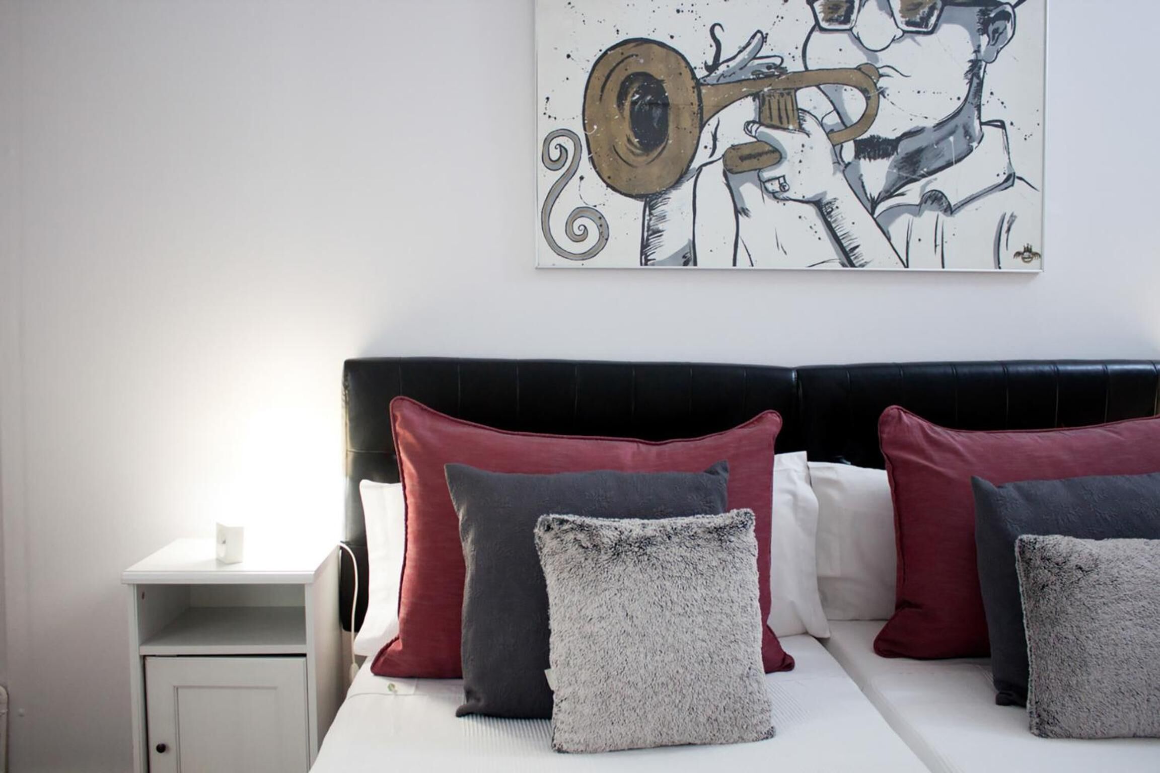 Un lit bien fait, avec une tête de lit noire, plusieurs oreillers rouges et gris, et une table de chevet blanche sur laquelle se trouve une lampe.