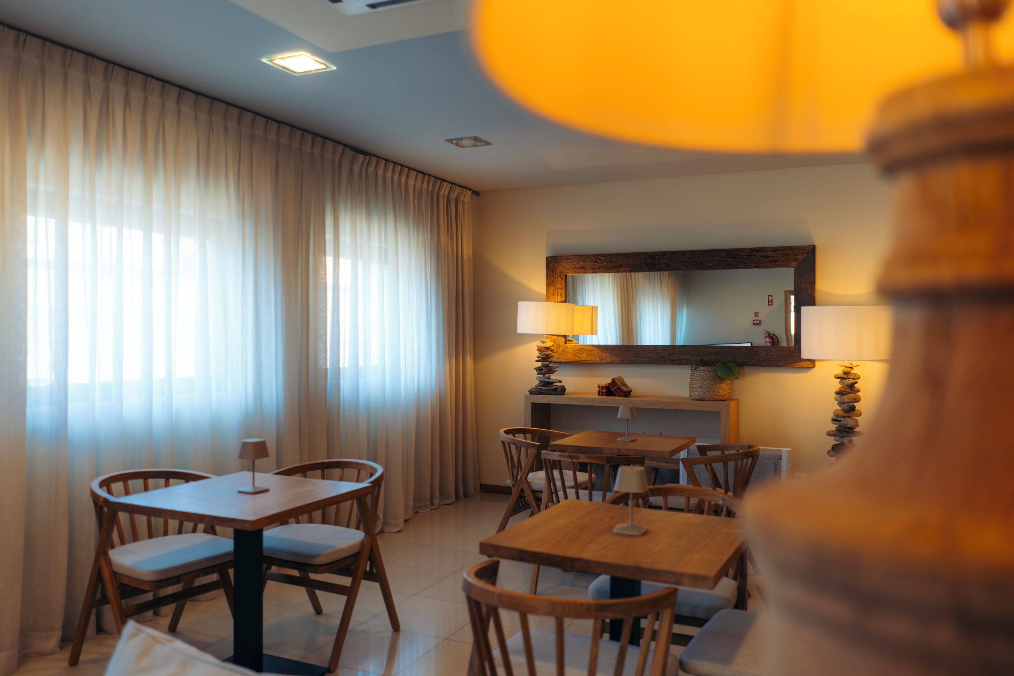 A dining area with wooden tables and chairs, a large window with curtains, and a mirror above the counter.
