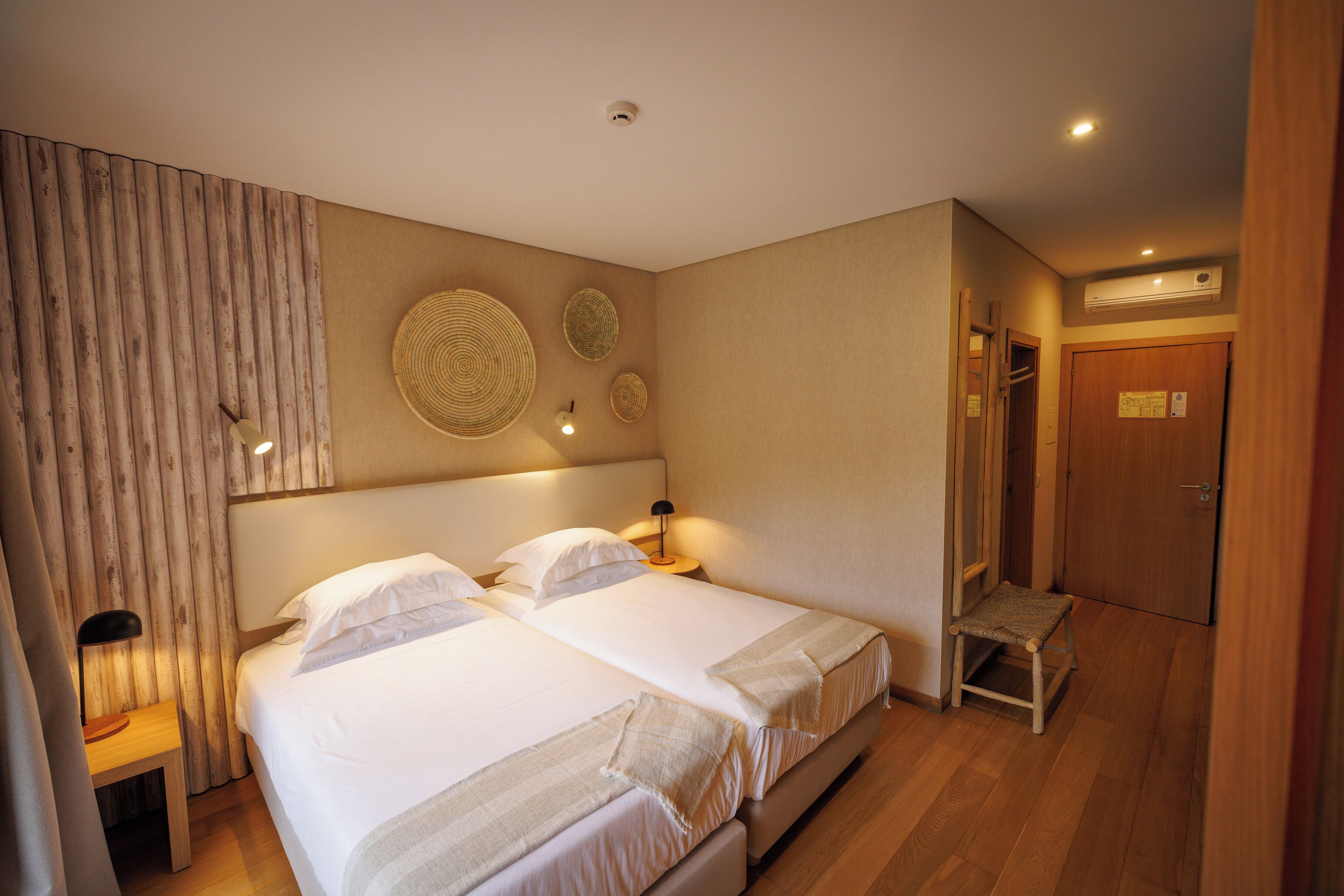 A hotel room with two beds, a wooden bench, a wall-mounted lamp, and a door with a towel rack.