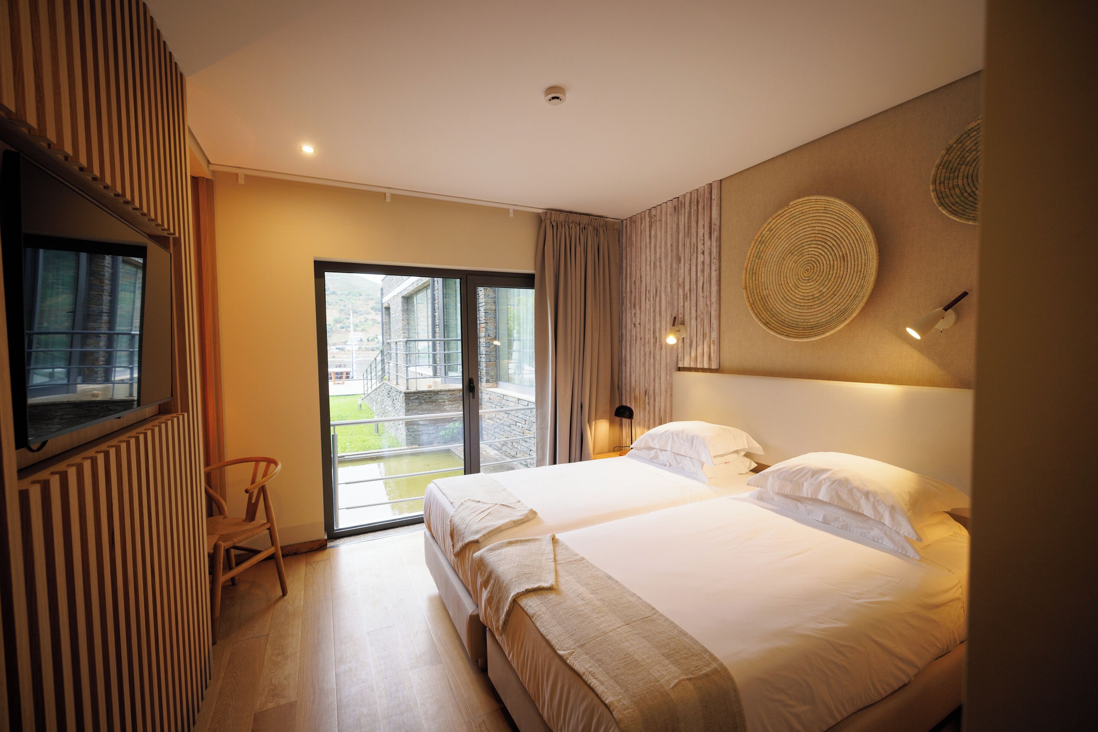 A modern hotel room with two beds, a large window, and a view of the outside.