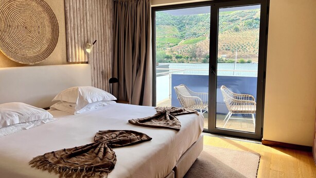 A hotel room with a large bed, a view of a hillside through a sliding door, and a balcony with chairs. - Hotel Folgosa Douro (Armamar)