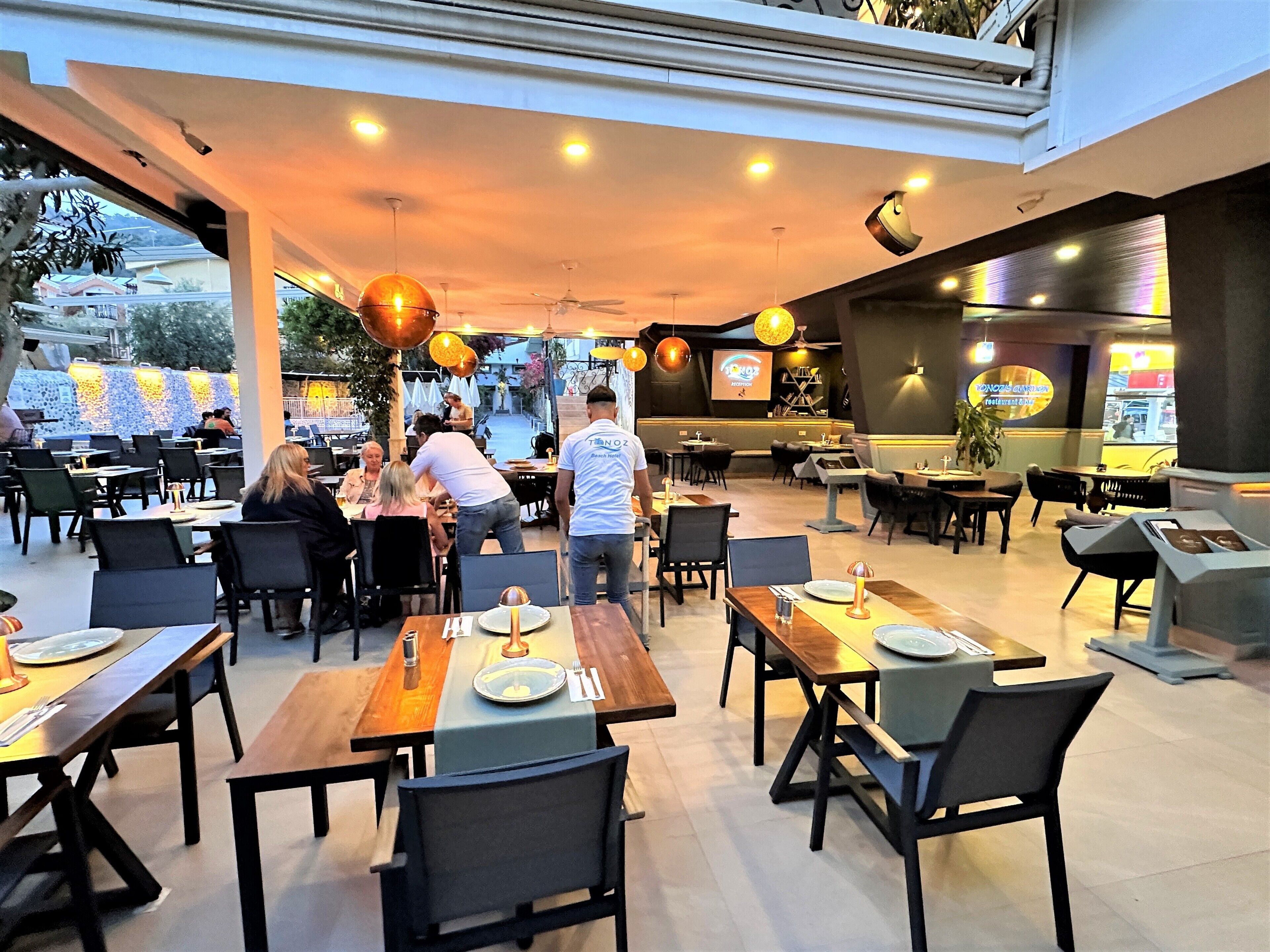 An indoor restaurant with multiple dining tables and chairs, a ceiling with hanging lights, and a view of the outside.