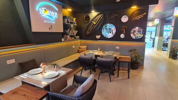 L’intérieur d’un restaurant, avec un espace d’accueil, des tables et des chaises disposées pour les clients, et un mur décoré d’instruments de musique.