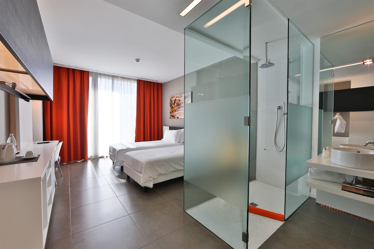 A modern hotel room with a glass-enclosed shower, a bed, a desk with a chair, and a large window with red curtains.