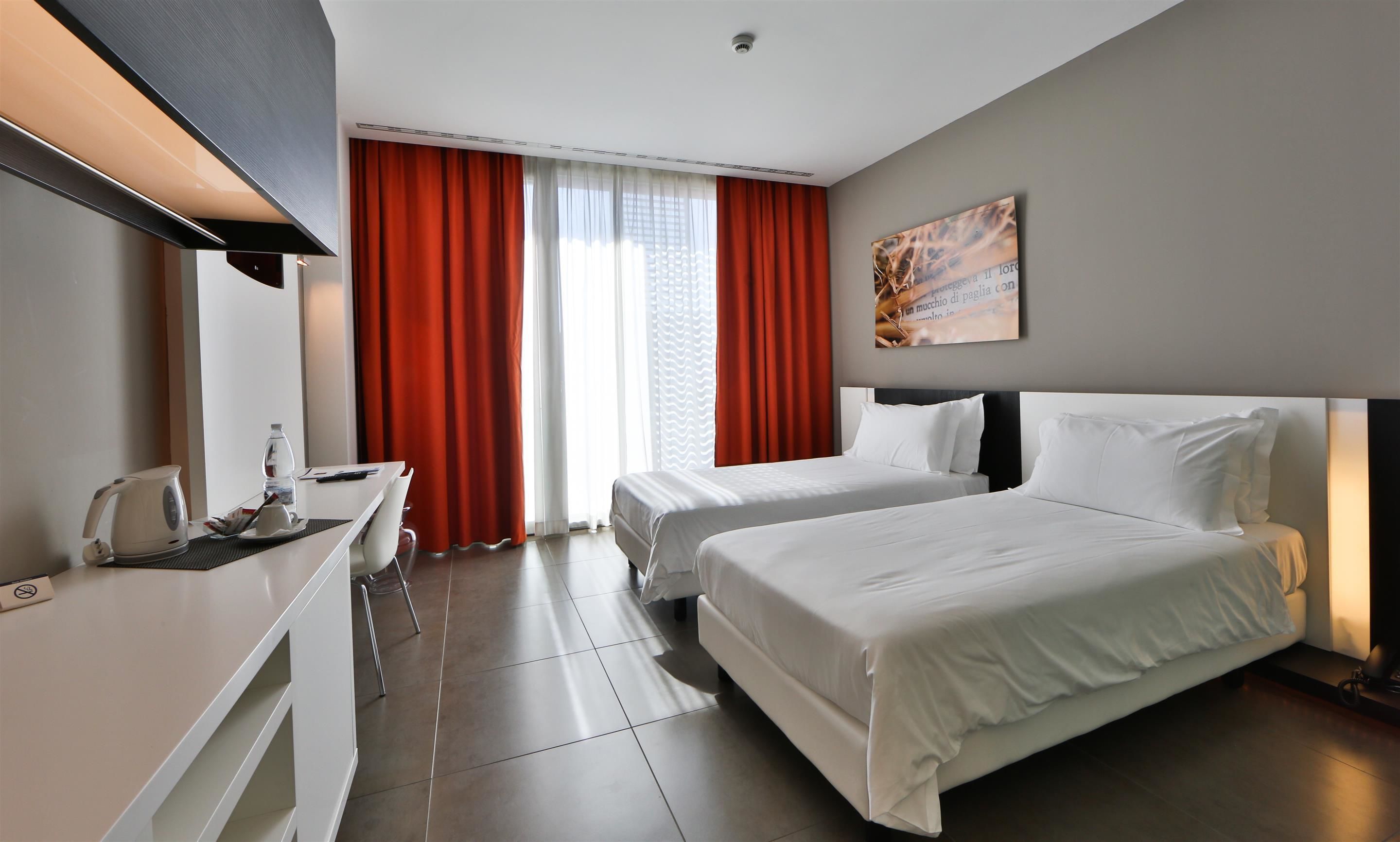 Habitación de hotel con dos camas, un escritorio, una tetera y una ventana con cortinas rojas.