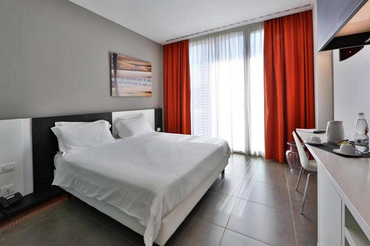A modern hotel room with a large bed, a desk with a chair, and a window with red curtains.