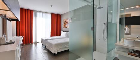 Habitación de hotel moderna con cama, zona de ducha con puertas de cristal, lavabo blanco y escritorio con silla.
