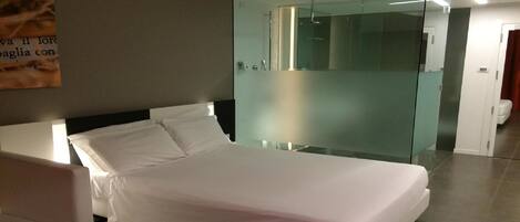 A modern hotel room with a bed, a glass partition, and a bathroom visible through the partition.