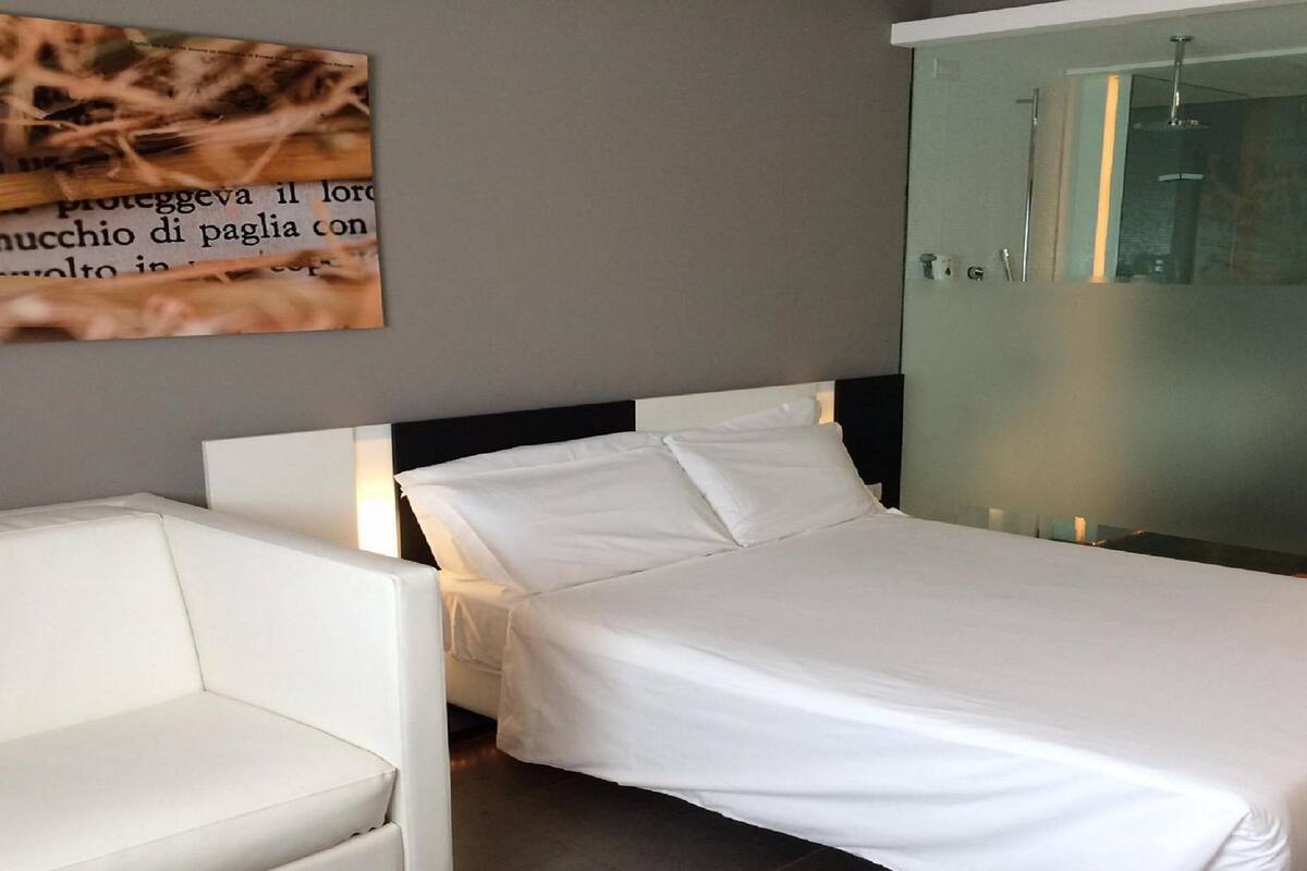 A modern bedroom with a bed, a white chair, and a glass shower enclosure.