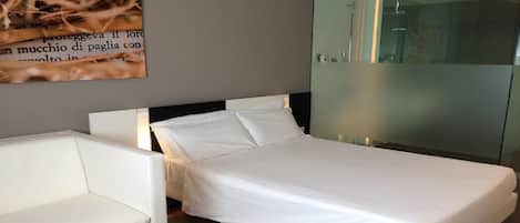 A modern bedroom with a bed, a white chair, and a glass shower enclosure.
