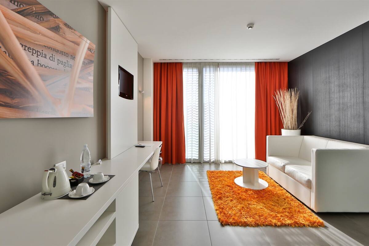 A modern hotel room with a white sofa, a coffee table, a TV mounted on the wall, and a large window with orange curtains.