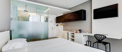 Una habitación de hotel moderna con una cama grande, un televisor montado en la pared, una pequeña kitchenette y un box de ducha de vidrio.