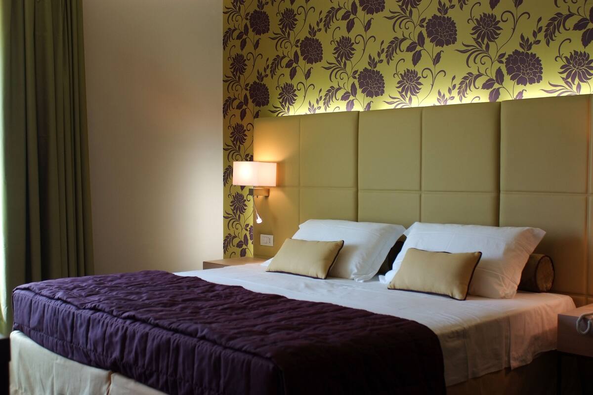 Una habitación de hotel con una cama grande, una mesita de noche con lámpara y papel tapiz con motivos florales.