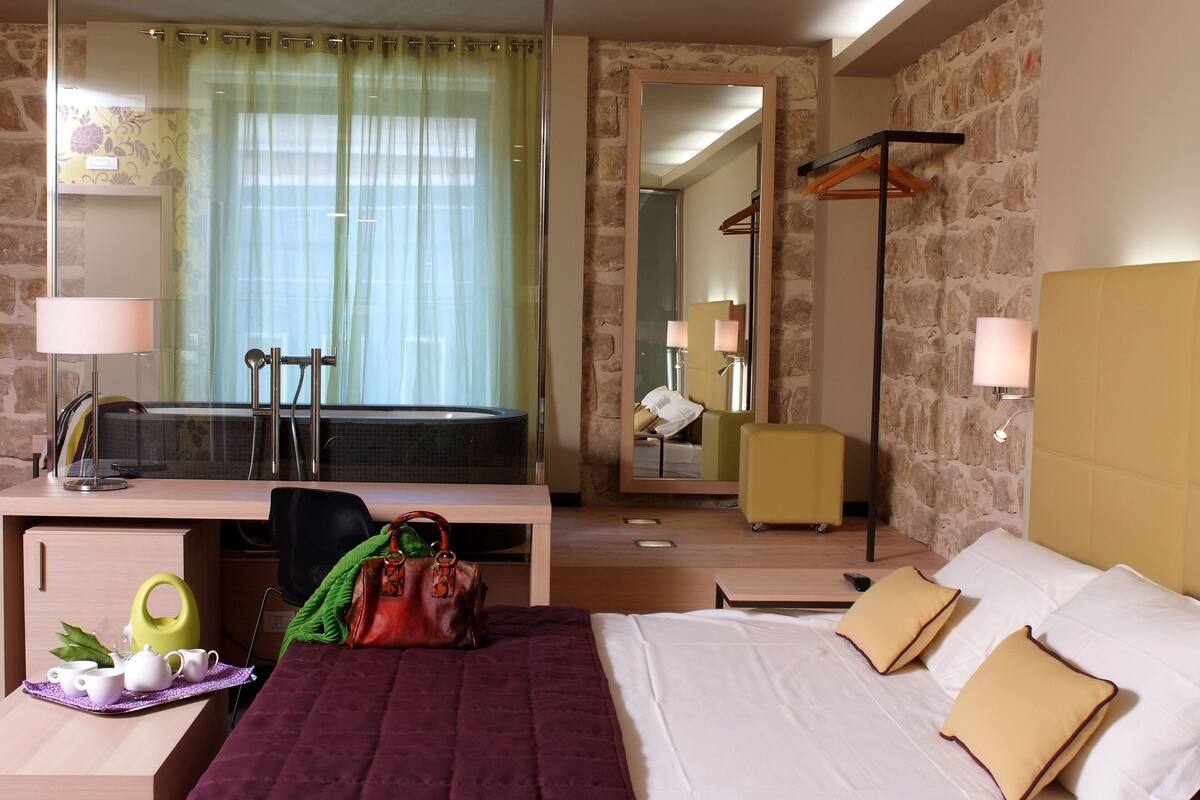 Habitación de hotel con cama, escritorio y baño visible a través de un separador de vidrio.