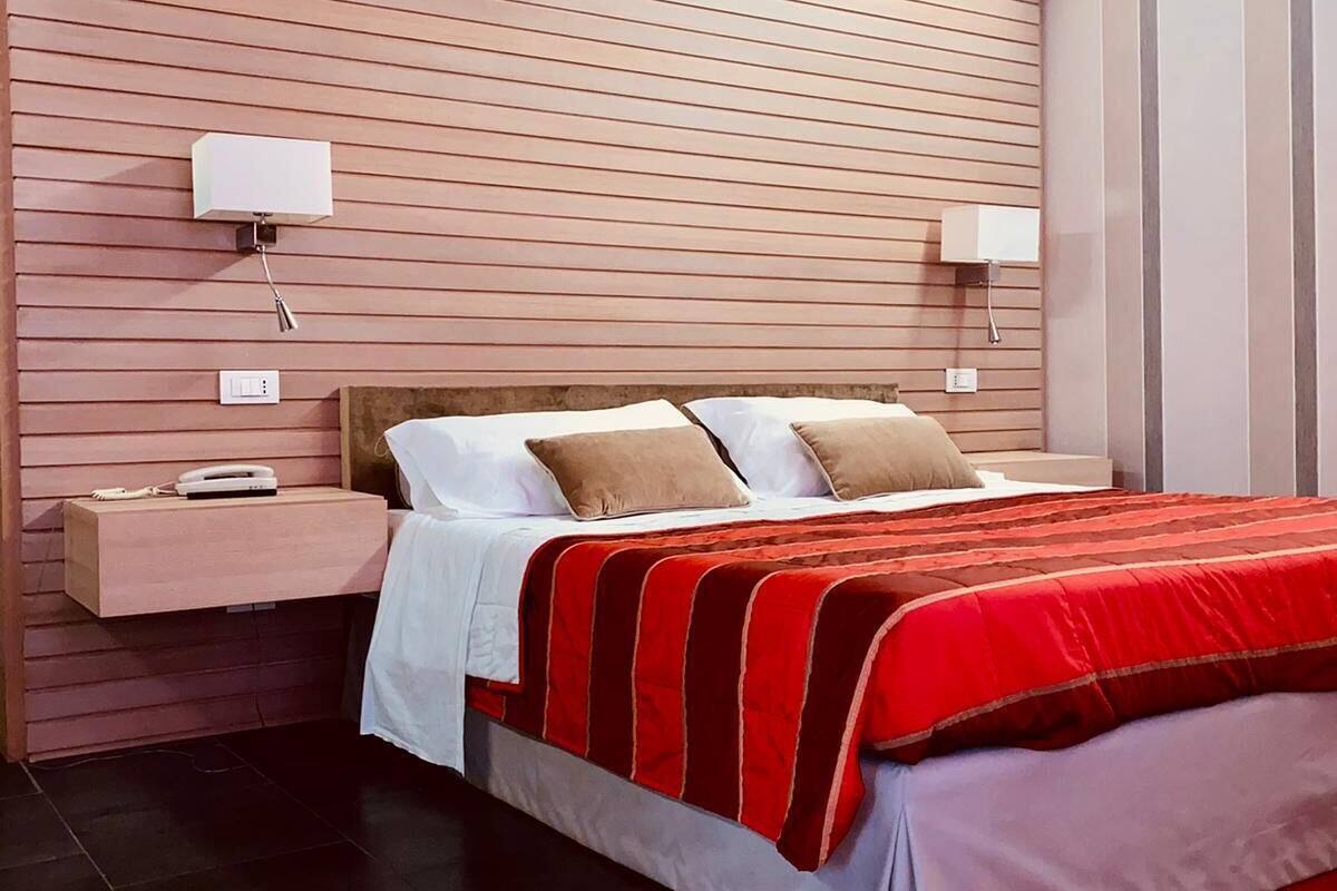 Un dormitorio moderno con pared de listones de madera, una cama grande con colcha a rayas rojas y blancas, dos mesitas de noche con lámparas y una alfombra roja en el suelo.