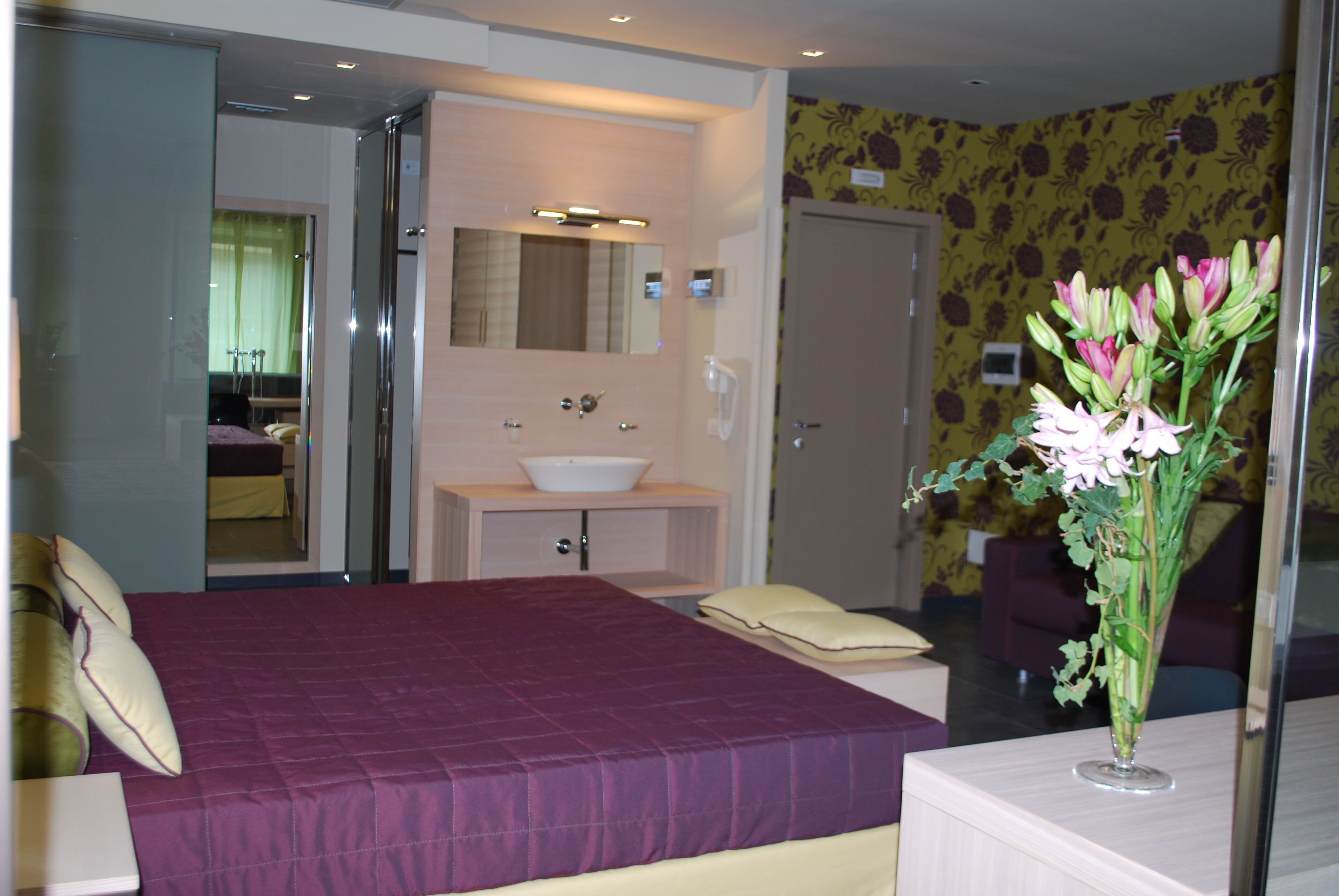 A hotel room with a bed, a bathroom with a sink and mirror, and a vase with flowers on a table.