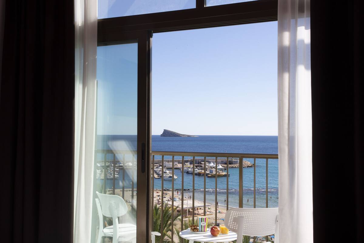 A balcony with a view of the sea, a table with a fruit bowl, and a chair.