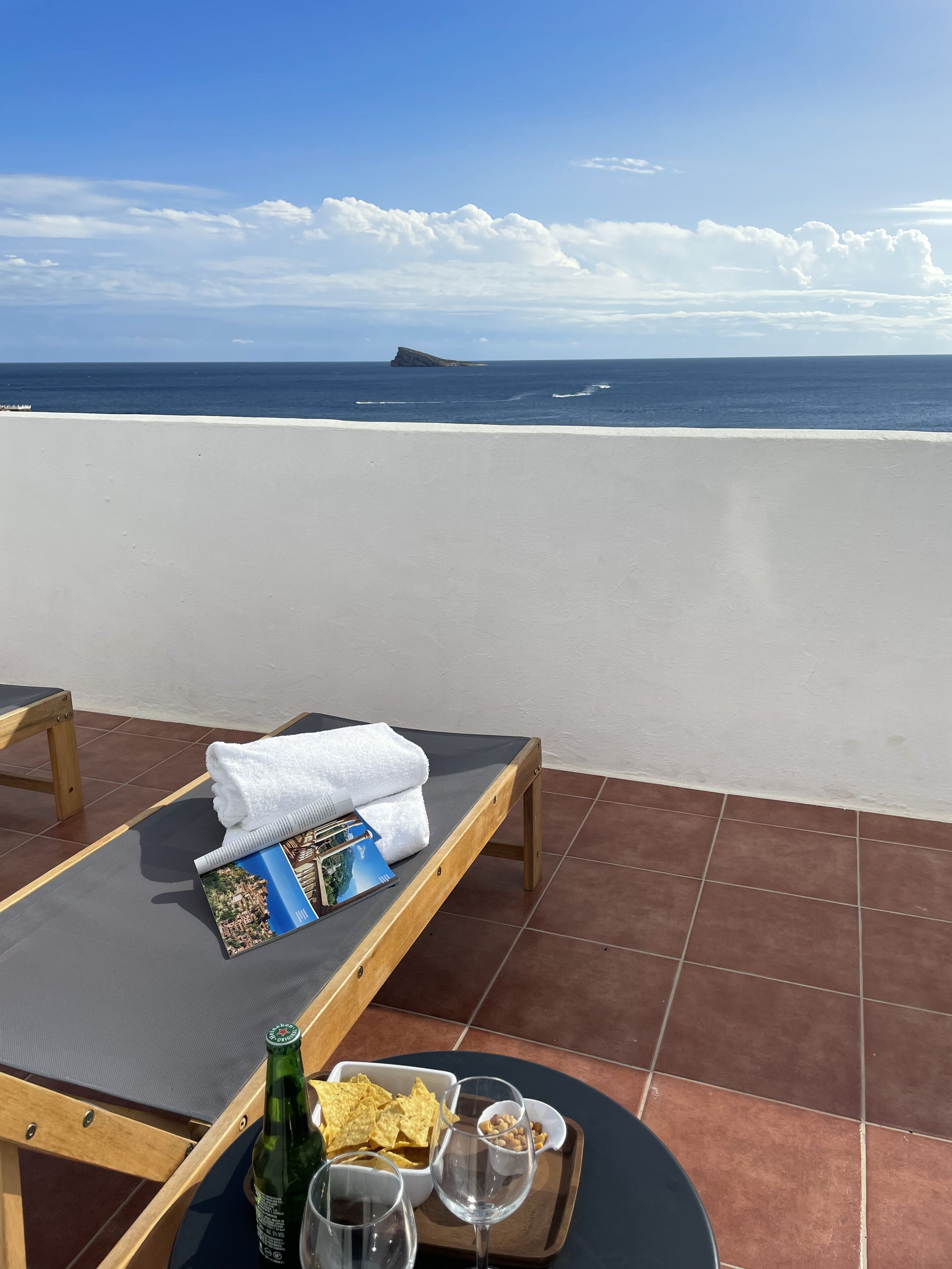 Una tumbona con una toalla y revistas, una bandeja con aperitivos y bebidas, y vistas al mar y una isla distante.