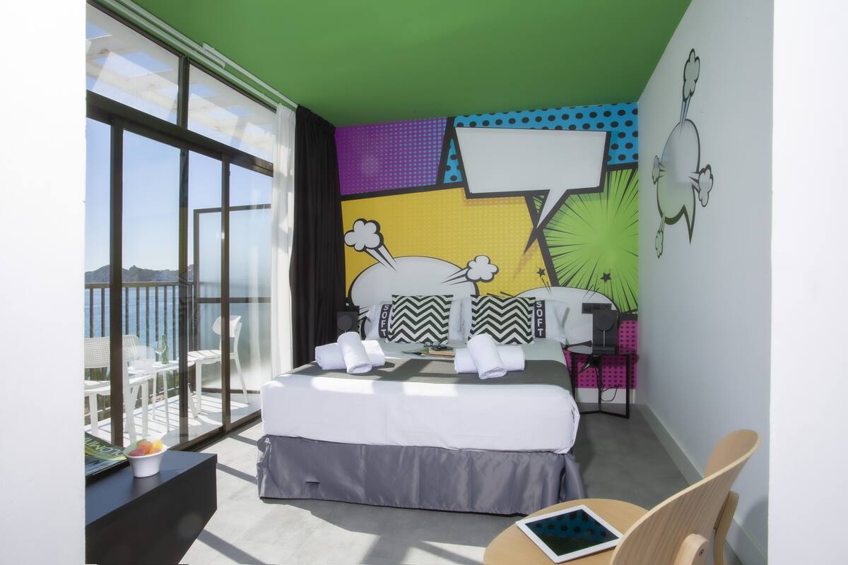 A modern hotel room with a large bed, a small desk, a chair, and a view of the outdoors.