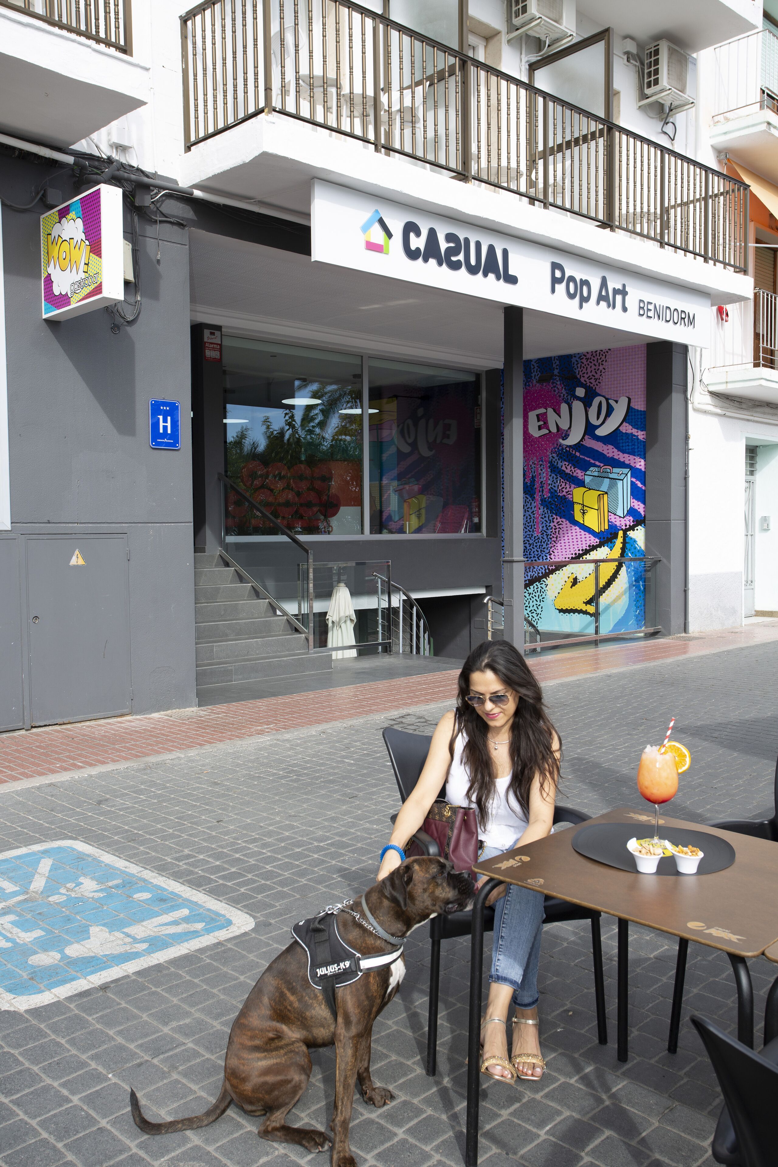 A woman sitting at an outdoor cafe with a dog, in front of a building with a 'Casual Pop Art' sign.