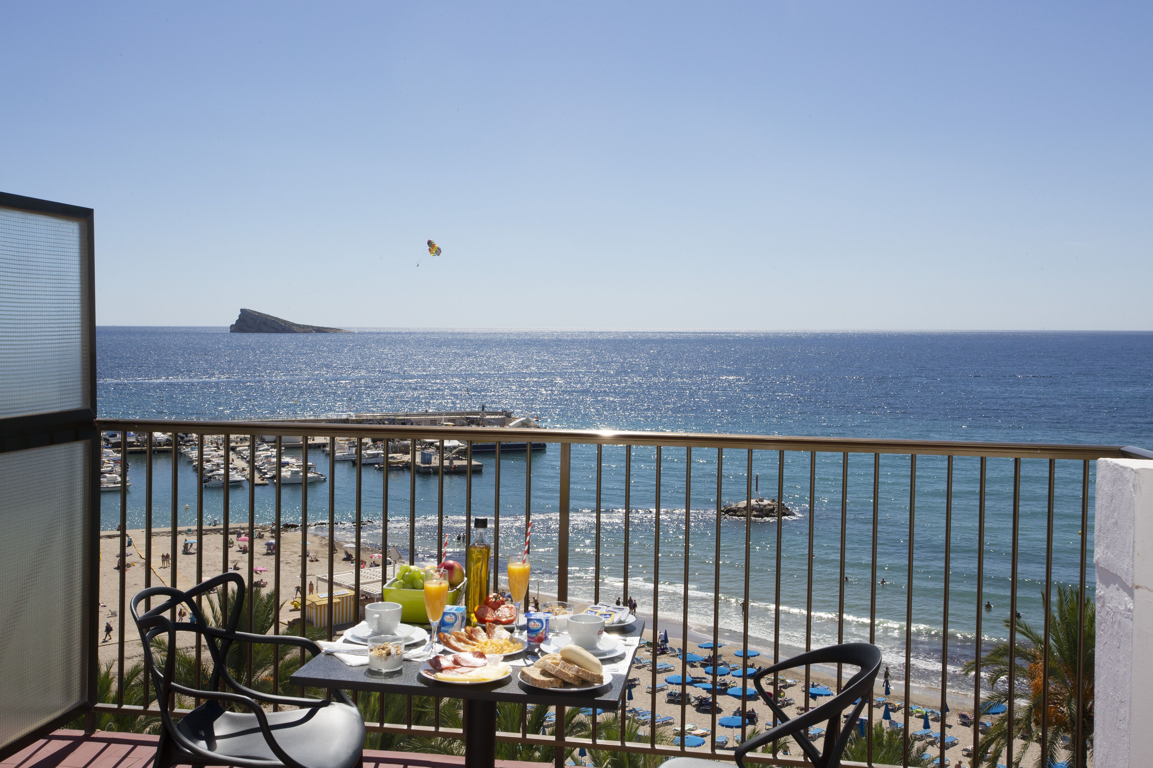 A balcony with a table set for breakfast overlooking a beach with boats and a clear sky.