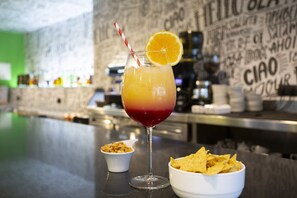 A cocktail with a red and yellow gradient, garnished with an orange slice and a striped straw, accompanied by a bowl of chips. - Casual Pop Art Hotel by Casual Hoteles (Benidorm)