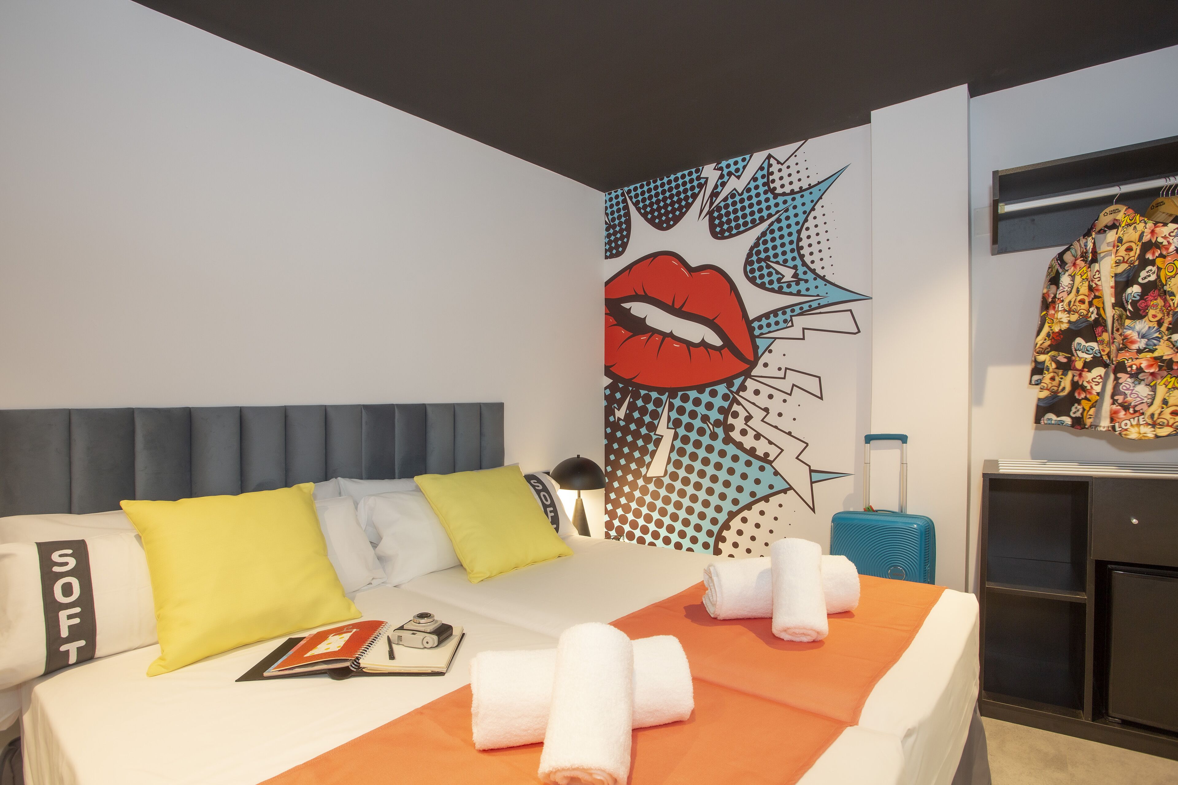 A hotel room with a bed, pillows, a wall art piece, a suitcase, and a wardrobe.
