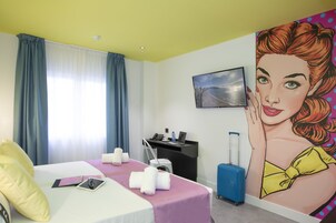 A hotel room with a bed, a desk with a laptop, a suitcase, and a painting on the wall.