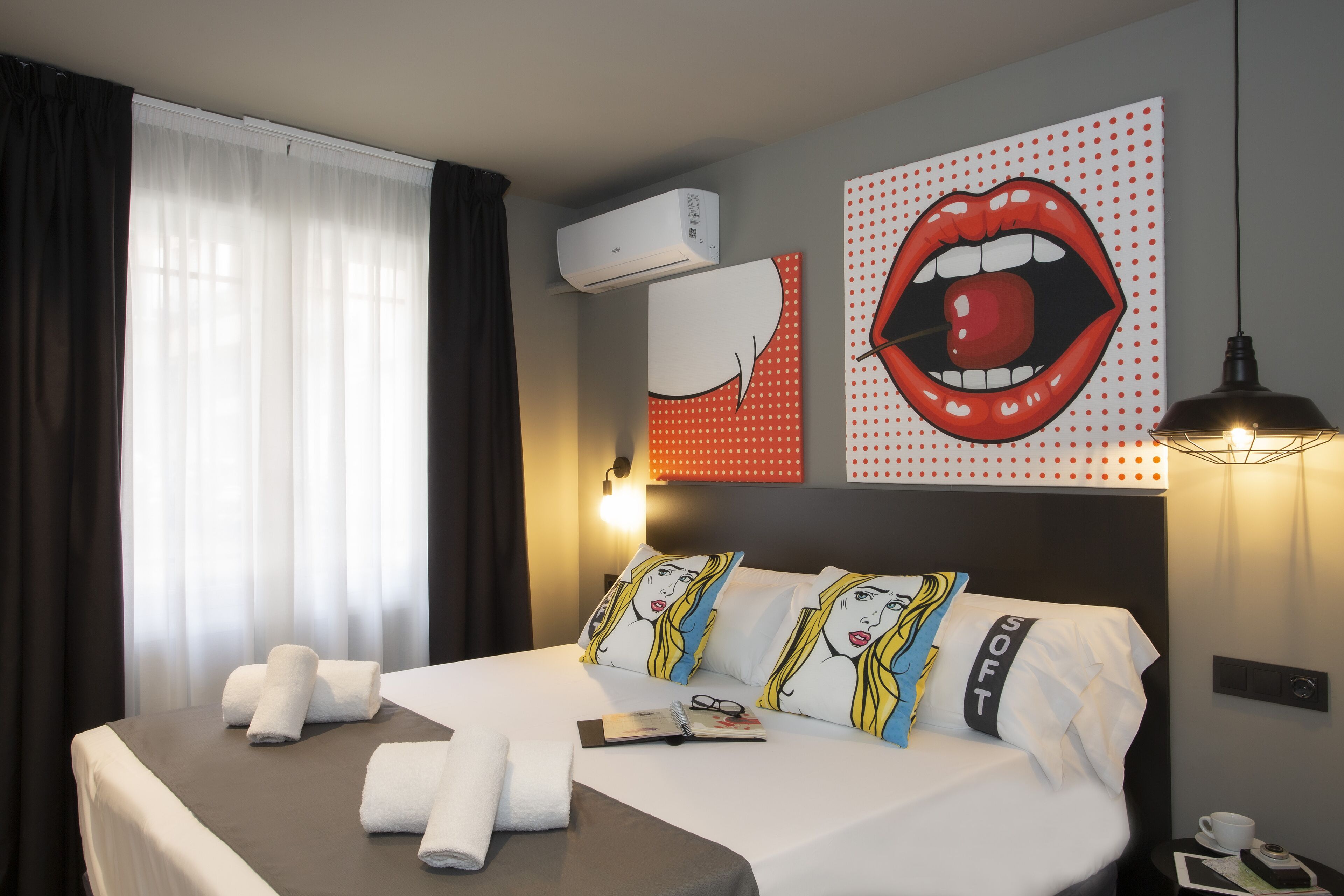 A hotel room with a bed, a window with curtains, an air conditioning unit, and a wall with a polka dot design.