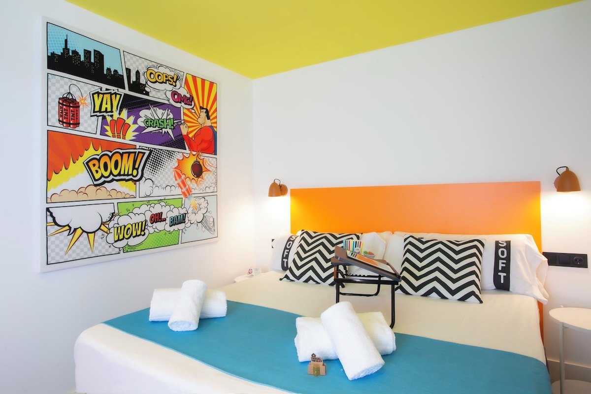 A hotel room with a bed, pillows, a headboard, a wall art comic strip, and a small table with a lamp.