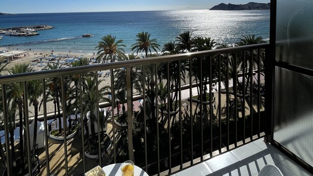 A balcony with a table, a glass of orange juice, and an apple. The view includes a beach, palm trees, and a clear blue sky.