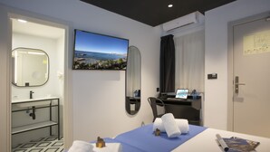 A hotel room with a bed, a desk, a chair, a TV, a bathroom, and a view of the sea.