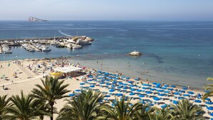 A beach with numerous blue umbrellas, a marina with boats, and a rocky outcrop in the distance.