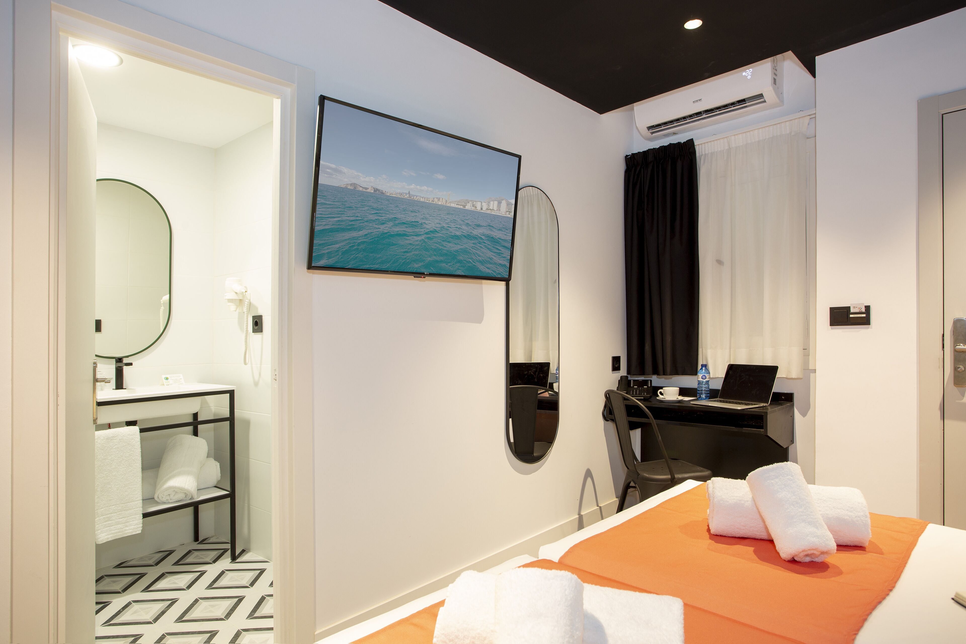 A hotel room with a bed, a desk with a laptop, a large window with a view of the sea, a bathroom with a mirror and towel rack, and an air conditioning unit.