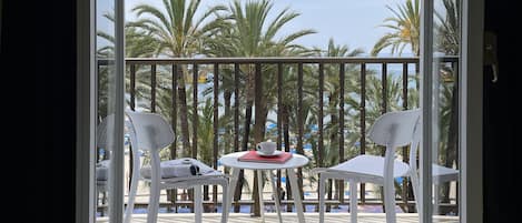Un balcón con vistas a palmeras y una playa. Hay sillas blancas y una pequeña mesa con una taza encima.