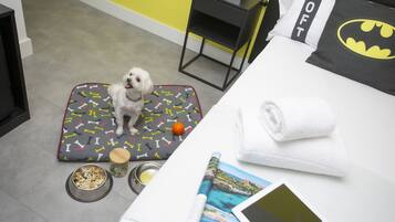 Una cama para mascotas con estampado de huesos, un tazón pequeño de comida para perro, un tazón de agua y una revista enrollada sobre una cama con un almohadón de Batman y una toalla blanca doblada.