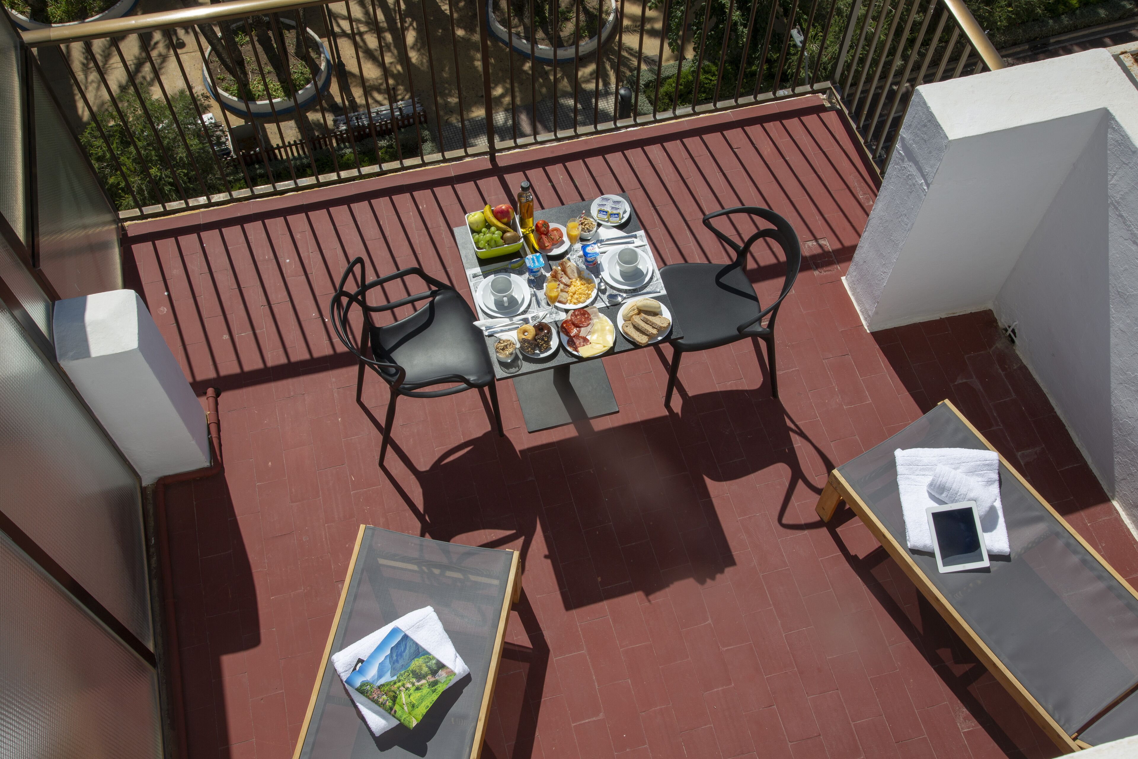 Un balcón con una mesa dispuesta para el desayuno, dos sillas y una mesita con un libro y una toalla.