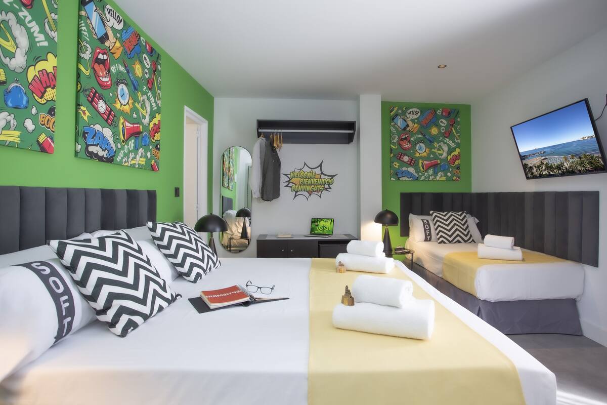 A modern hotel room with a large bed, a sofa, and a desk. There are green walls and a large window with a view of the beach.