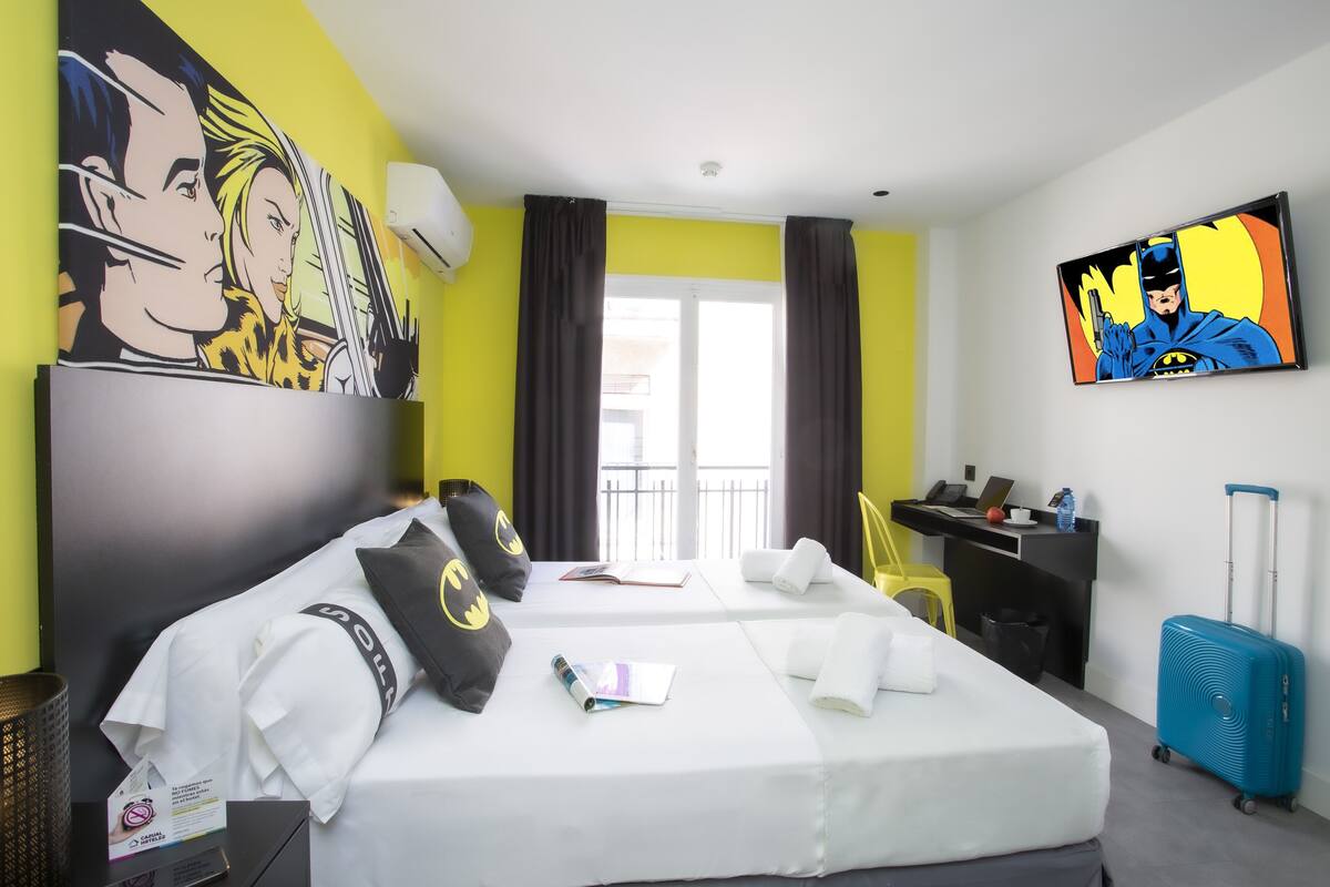 A hotel room with two beds, a desk, a chair, a suitcase, and artwork on the walls.
