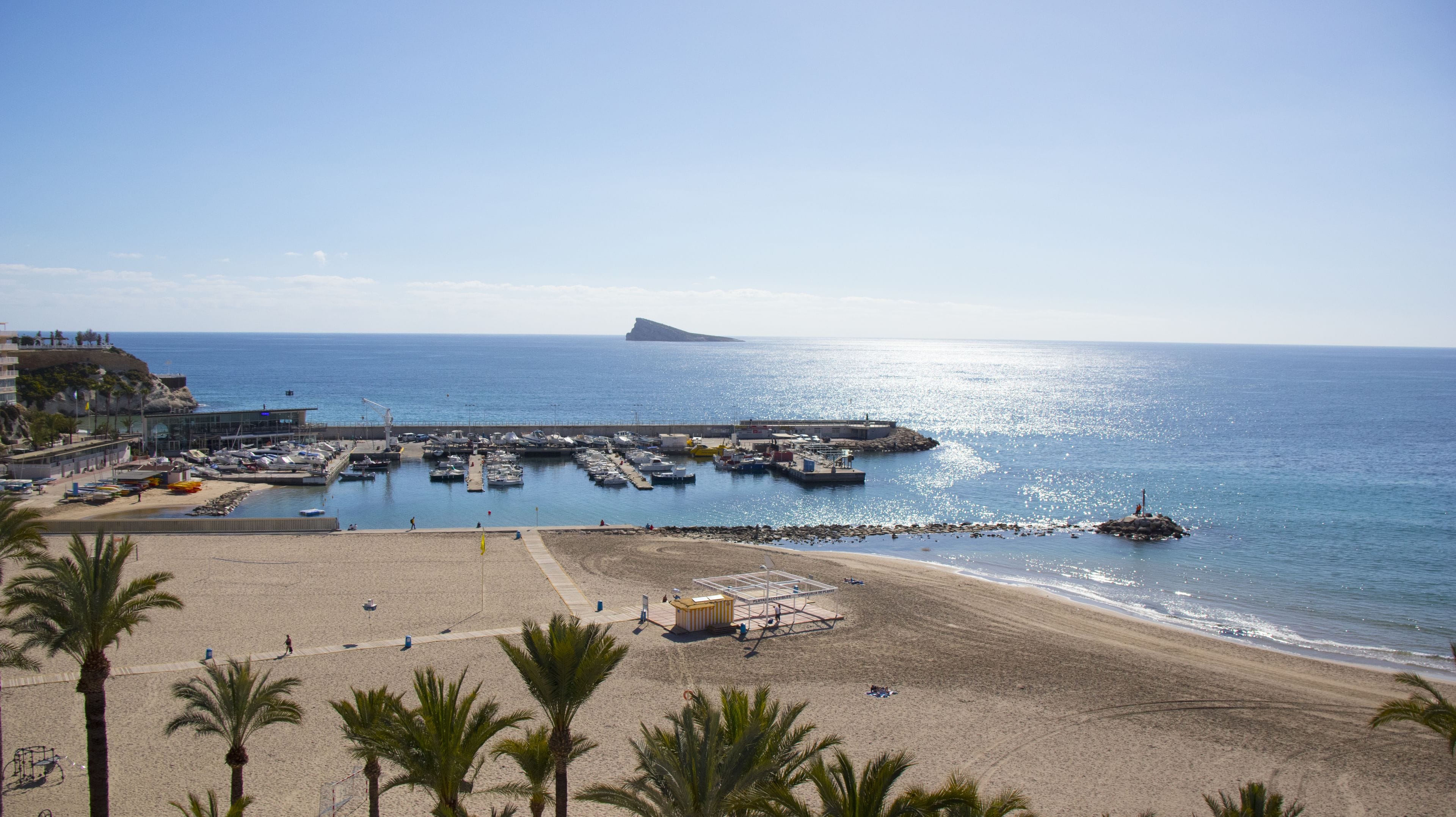 A coastal beach with a marina, palm trees, and a rocky outcrop in the distance.